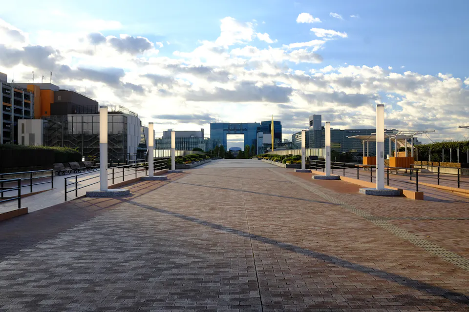 A wide, paved walkway bordered by pillars extends towards a modern urban complex in the distance. Fluffy clouds fill the blue sky above, with sunlit shadows creating patterns on the ground.