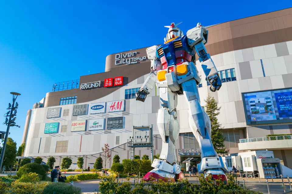 A giant Gundam statue stands in front of the DiverCity Tokyo Plaza. The building features various brand logos, including Uniqlo and H&M, against a clear blue sky. People are gathered around the statue, taking photos.