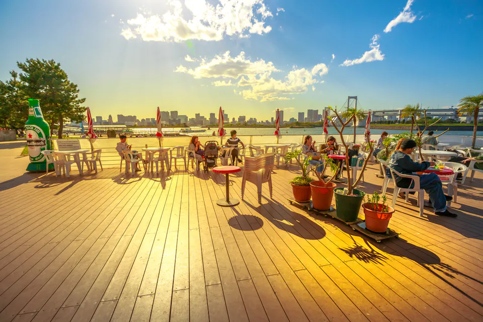 A sunny outdoor deck with tables and chairs overlooks a city skyline. A few people relax under umbrellas. Potted plants line the area, and a large bottle decoration stands nearby. The sky is bright with scattered clouds.