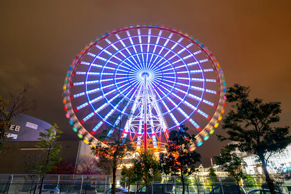 A brightly lit Ferris wheel spins at night, displaying colorful light patterns against a cloudy sky. Trees and buildings are visible in the foreground, adding depth to the vibrant scene.