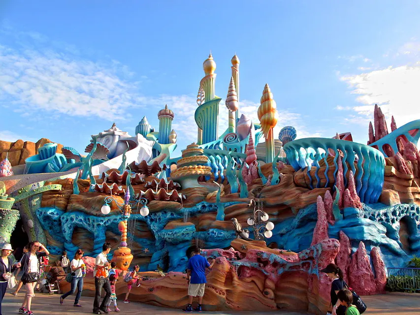 Colorful, whimsical theme park scene with ornate, fantasy-style architecture featuring seashells and coral. People walk around enjoying the atmosphere under a bright blue sky.
