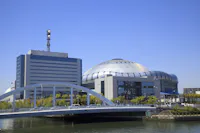 A modern white bridge crosses a river in front of a large, dome-shaped stadium and an adjacent office building, both under a clear blue sky. Trees line the riverbank.