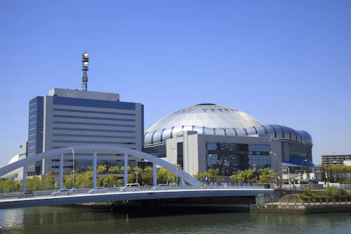 A modern white bridge crosses a river in front of a large, dome-shaped stadium and an adjacent office building, both under a clear blue sky. Trees line the riverbank.