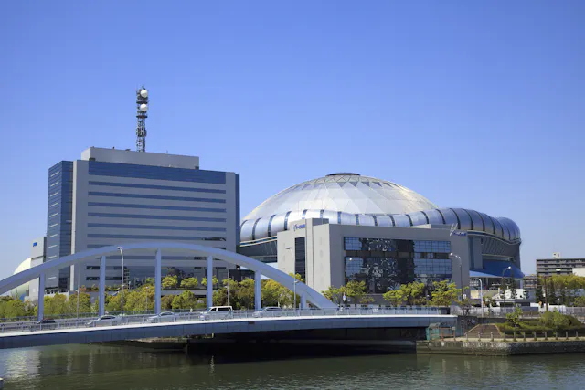 A modern white bridge crosses a river in front of a large, dome-shaped stadium and an adjacent office building, both under a clear blue sky. Trees line the riverbank.
