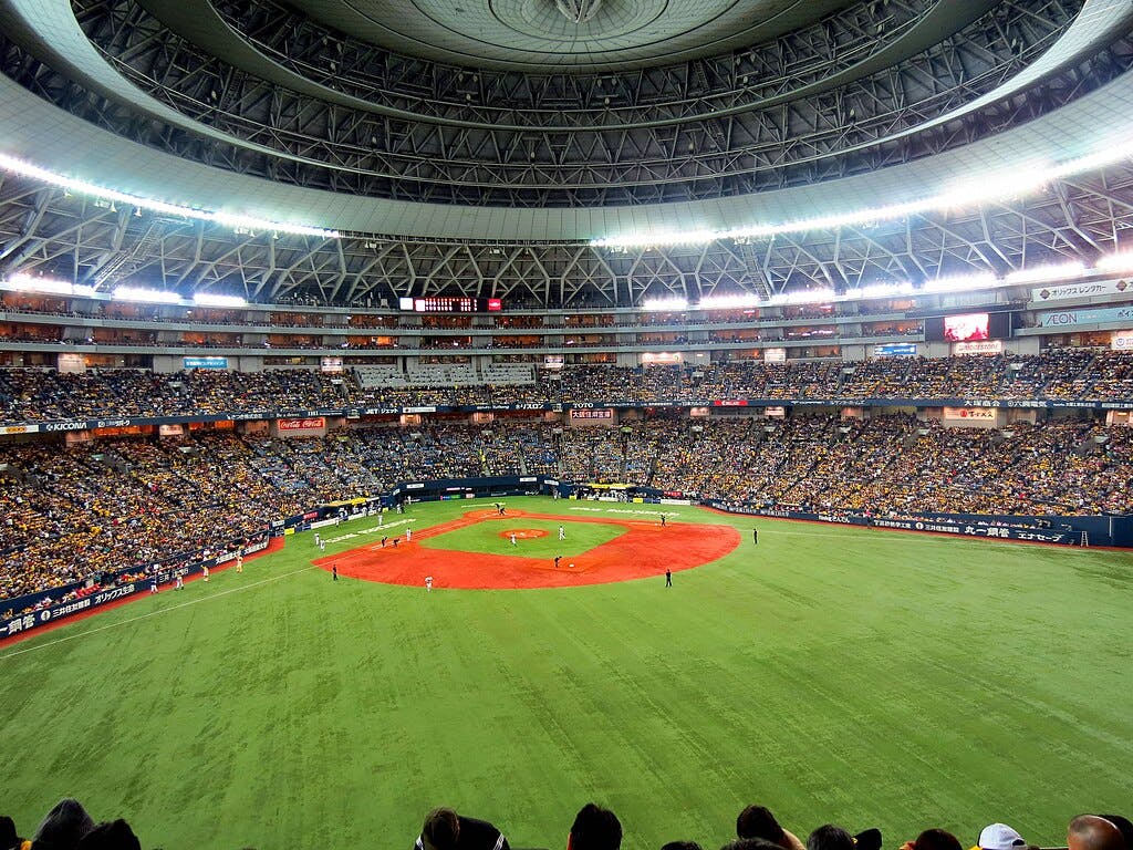 A large indoor baseball stadium filled with spectators, with players on the bright green field and a domed, intricately structured roof overhead. The stands are packed, and advertisements line the walls above the crowds.