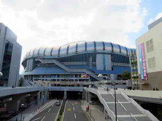 A large, modern stadium with a curved, silver and blue exterior stands beneath a partly cloudy sky. Surrounding buildings and elevated walkways lead to its main entrance, and a few people and vehicles are visible below.