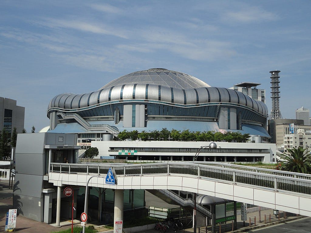 A large, modern stadium with a silver domed roof and curved design, surrounded by urban structures and a pedestrian bridge in the foreground under a blue sky.