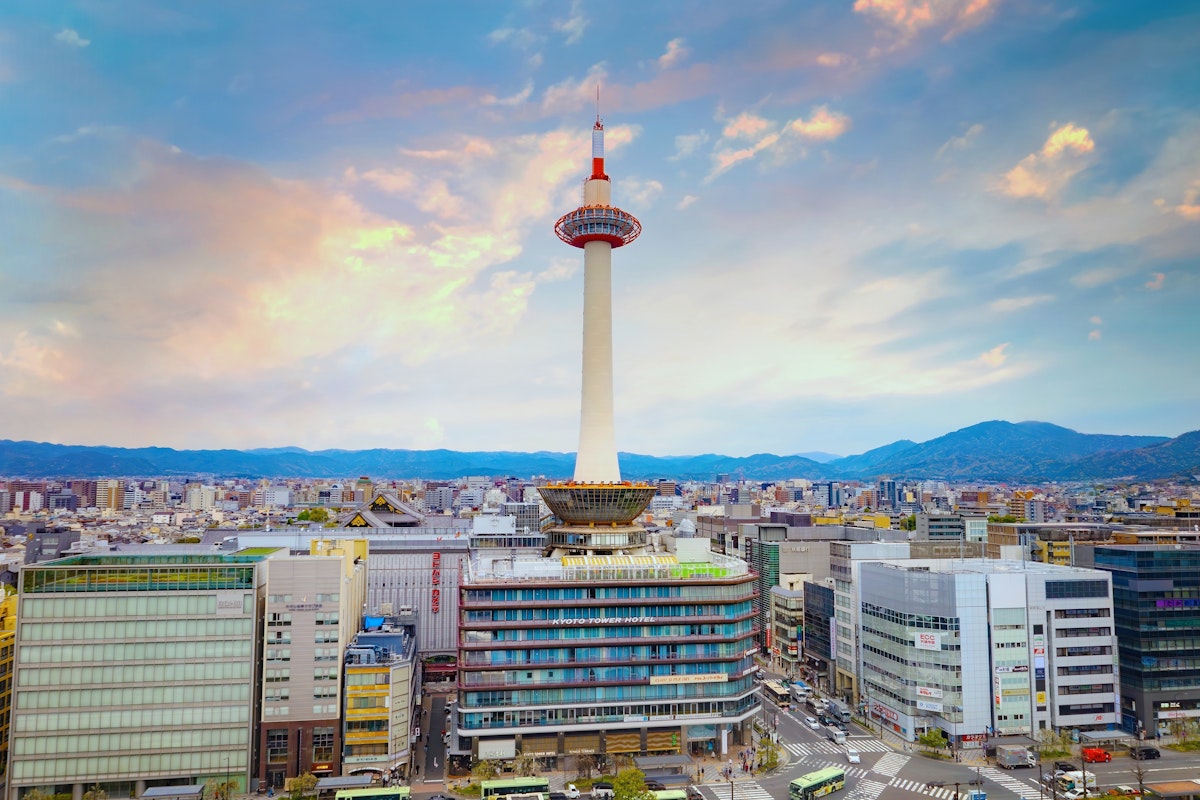 Kyoto Tower A cityscape at sunset featuring a tall white tower with a red observation deck. Surrounding the tower are modern buildings, and mountains can be seen in the background under a sky with colorful clouds.