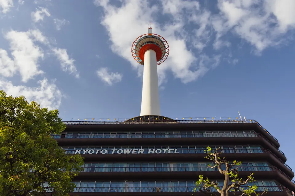 View of Kyoto Tower against a clear blue sky with scattered clouds. The Kyoto Tower Hotel is visible at the base, and a tree with green leaves is in the foreground.