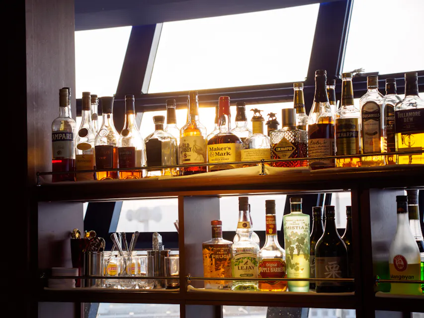 A backlit bar shelf displays various bottles of liquor, including whiskey and rum. Sunlight filters through a window behind the bottles, casting a warm glow. The shelf also has some glasses and a few decorative items.