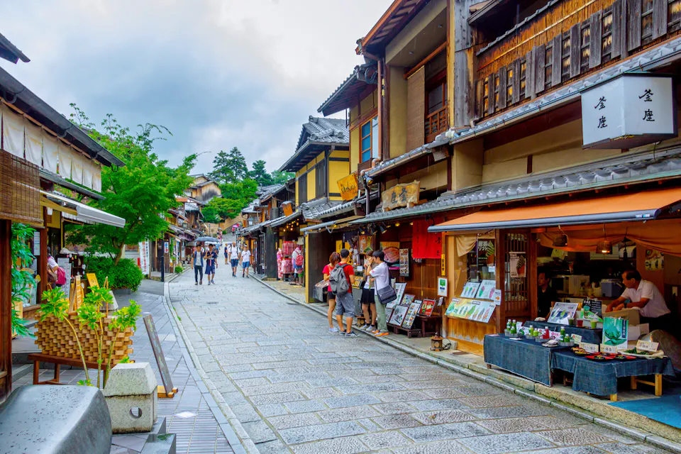 A vibrant street in a traditional Japanese town, lined with wooden buildings and shops displaying various goods. People walk along the stone pavement, and green trees can be seen in the background under a partly cloudy sky.