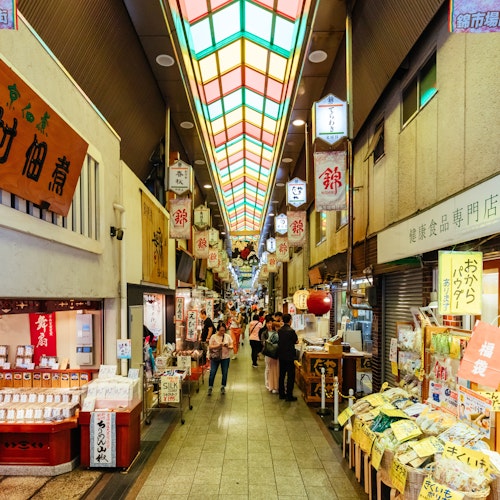 Nishiki Market Bustling indoor Japanese market scene with shops on both sides, colorful signs, and a stained glass ceiling. Shoppers explore stores displaying various goods, including food and souvenirs. Warm lighting adds to the lively atmosphere.