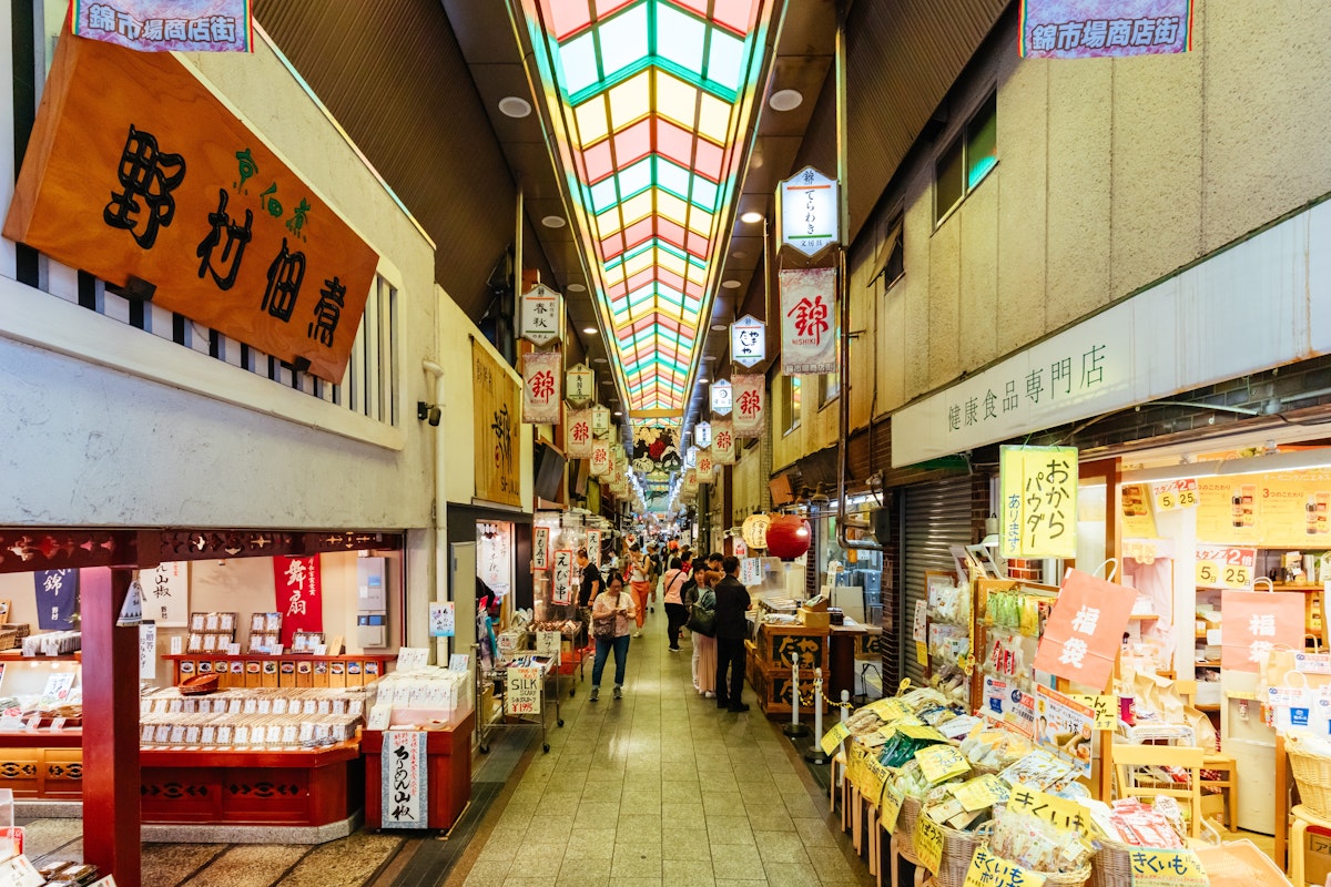 Nishiki Market Bustling indoor Japanese market scene with shops on both sides, colorful signs, and a stained glass ceiling. Shoppers explore stores displaying various goods, including food and souvenirs. Warm lighting adds to the lively atmosphere.