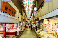 Bustling indoor Japanese market scene with shops on both sides, colorful signs, and a stained glass ceiling. Shoppers explore stores displaying various goods, including food and souvenirs. Warm lighting adds to the lively atmosphere.