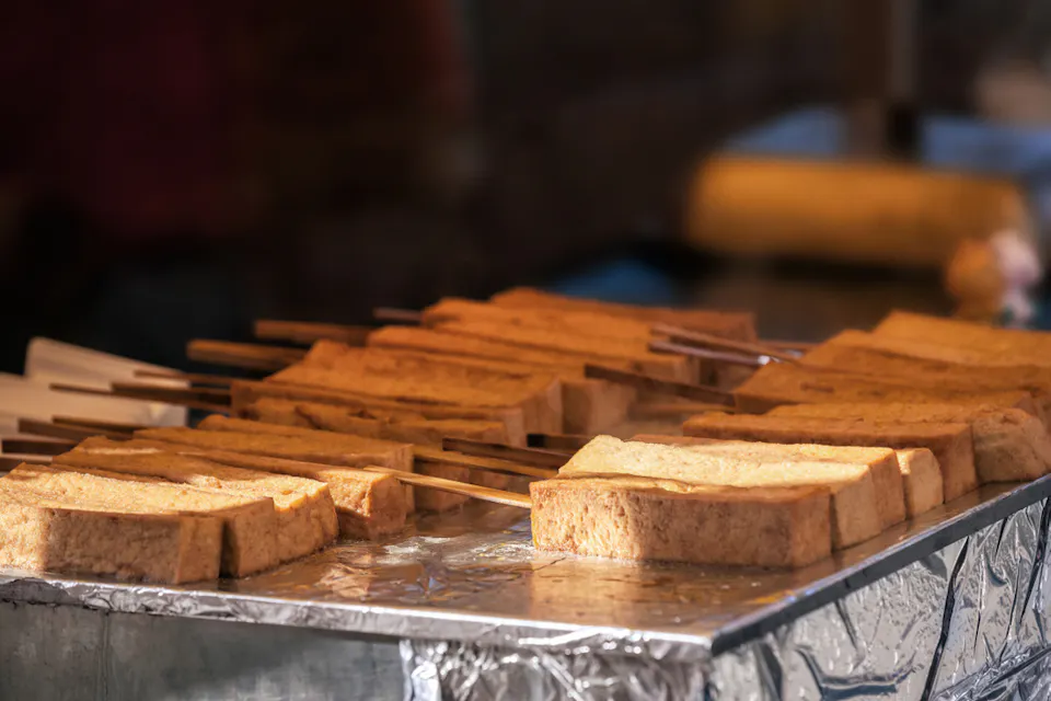 Skewers of fried tofu are lined up on a metallic surface, ready for cooking. The tofu looks golden brown and is arranged neatly, with chopsticks inserted into each piece. The background is softly blurred.