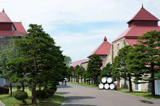 A quiet path lined with neatly trimmed trees runs between stone buildings with red roofs. Several white barrels are stacked on the right, and a few people walk in the distance under a partly cloudy sky.