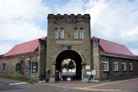 A stone building with a central archway entrance, red roofs, and rectangular windows. People are walking through the arch, and signs are posted near the entrance. The sky is partly cloudy.