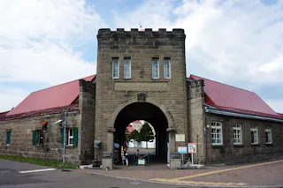 A stone building with a central archway entrance, red roofs, and rectangular windows. People are walking through the arch, and signs are posted near the entrance. The sky is partly cloudy.
