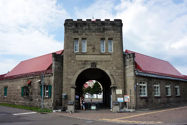 A stone building with a central archway entrance, red roofs, and rectangular windows. People are walking through the arch, and signs are posted near the entrance. The sky is partly cloudy.