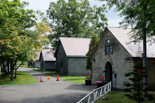Pathway lined with trees curves past three stone buildings with gabled roofs. Orange traffic cones and a barrier partially block the path. Green foliage and potted flowers are visible by the entrance.