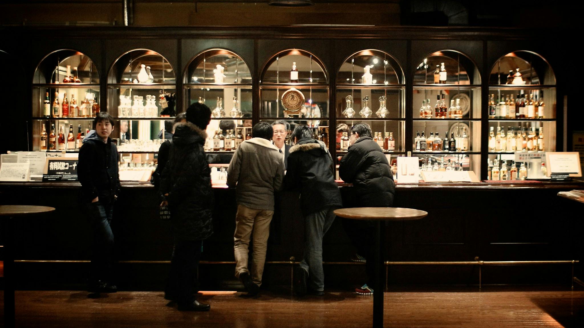 Five people in winter coats stand at a dimly lit bar with shelves of various bottles behind a dark wooden counter. The setting is warm, with drinks and menus visible on the bar.