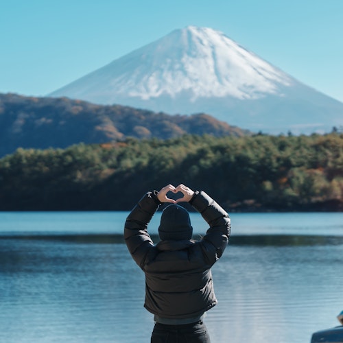 Mount Fuji Day Trip A person in a black jacket stands by a lake making a heart shape with their hands, facing Mount Fuji. The mountain is snow-capped and surrounded by clear skies, with trees and autumn foliage around the lake.