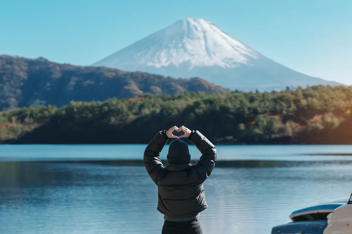 Mount Fuji Day Trip A person in a black jacket stands by a lake making a heart shape with their hands, facing Mount Fuji. The mountain is snow-capped and surrounded by clear skies, with trees and autumn foliage around the lake.
