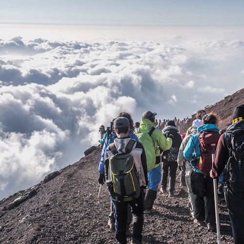 Outdoor Adventures Around Mount Fuji A group of hikers descends a rocky mountain trail surrounded by clouds. The scene depicts a high-altitude adventure with people wearing backpacks and outdoor gear, enjoying a spectacular view above the clouds.