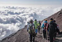 A group of hikers descends a rocky mountain trail surrounded by clouds. The scene depicts a high-altitude adventure with people wearing backpacks and outdoor gear, enjoying a spectacular view above the clouds.