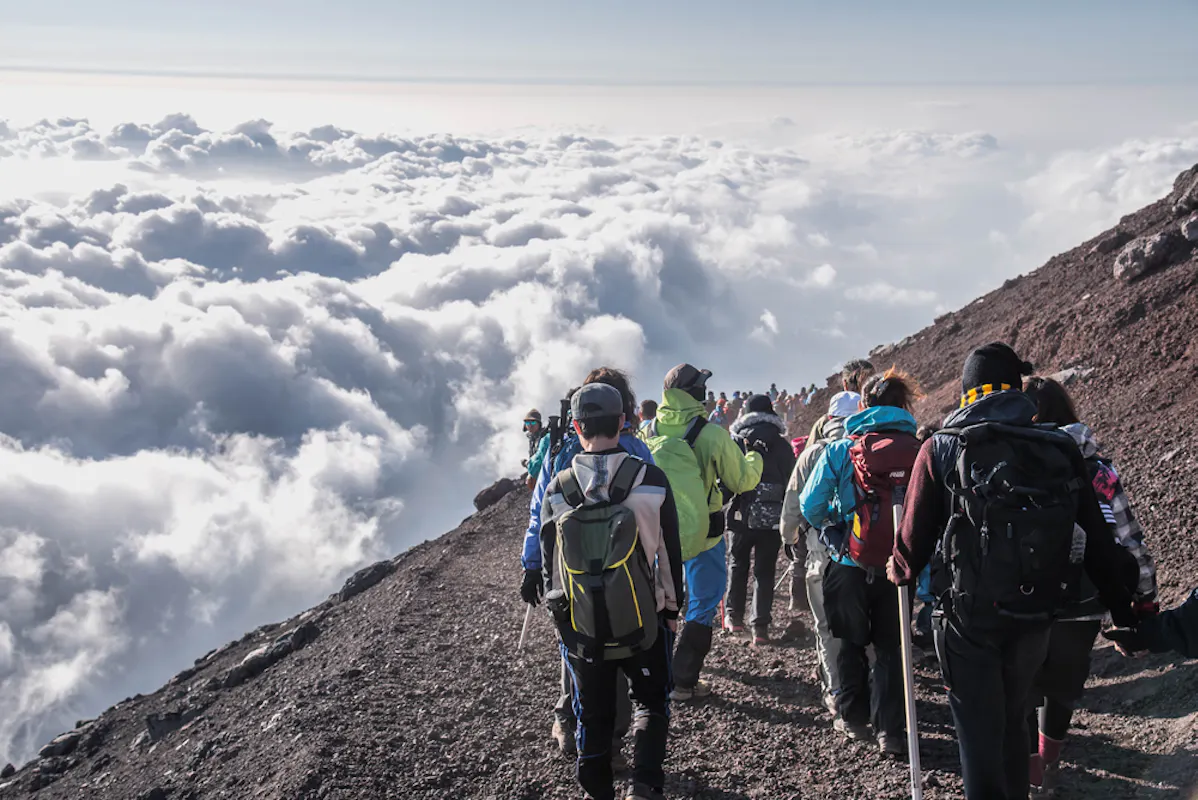 A group of hikers descends a rocky mountain trail surrounded by clouds. The scene depicts a high-altitude adventure with people wearing backpacks and outdoor gear, enjoying a spectacular view above the clouds.