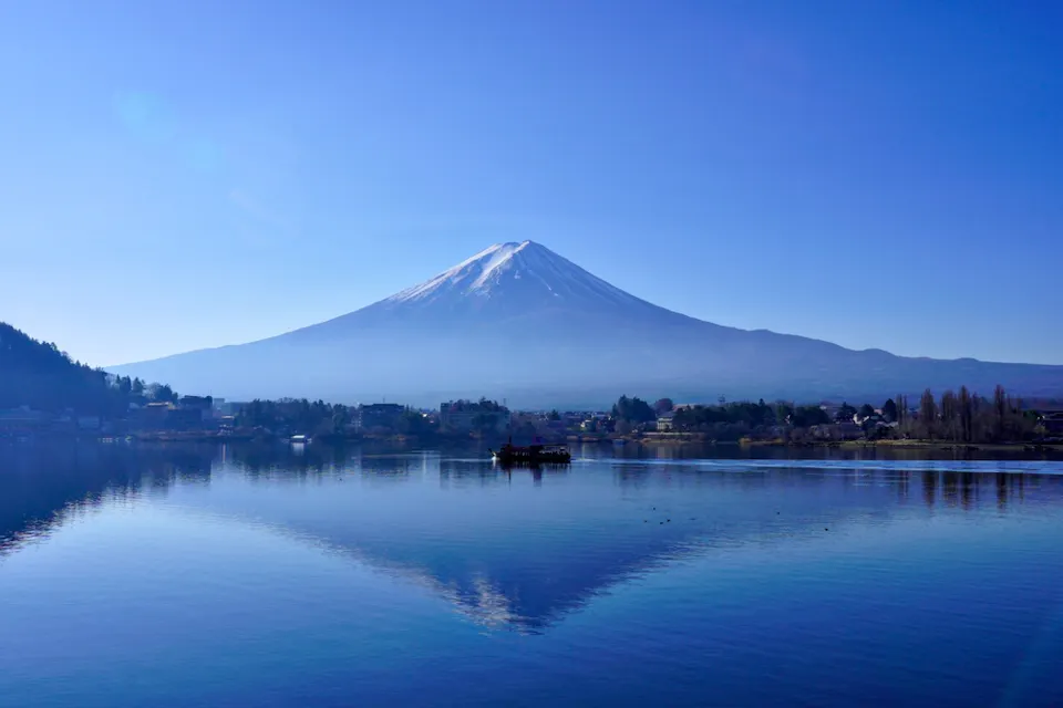 Taking in the Scenery A serene view of Mount Fuji with a clear blue sky, reflecting perfectly on a calm lake. Small buildings and trees line the shore, adding to the tranquility of the scene.