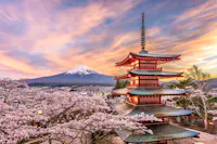 A five-story pagoda stands amid cherry blossoms, with Mount Fuji in the background under a vibrant sunset sky. The scene captures the serene beauty of the Japanese landscape in spring.