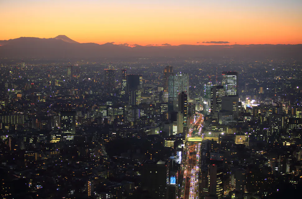 A city skyline at dusk with a vibrant sunset. Twinkling city lights illuminate the tall buildings, and a faint outline of a mountain, possibly Mount Fuji, is visible in the distance under an orange and purple sky.