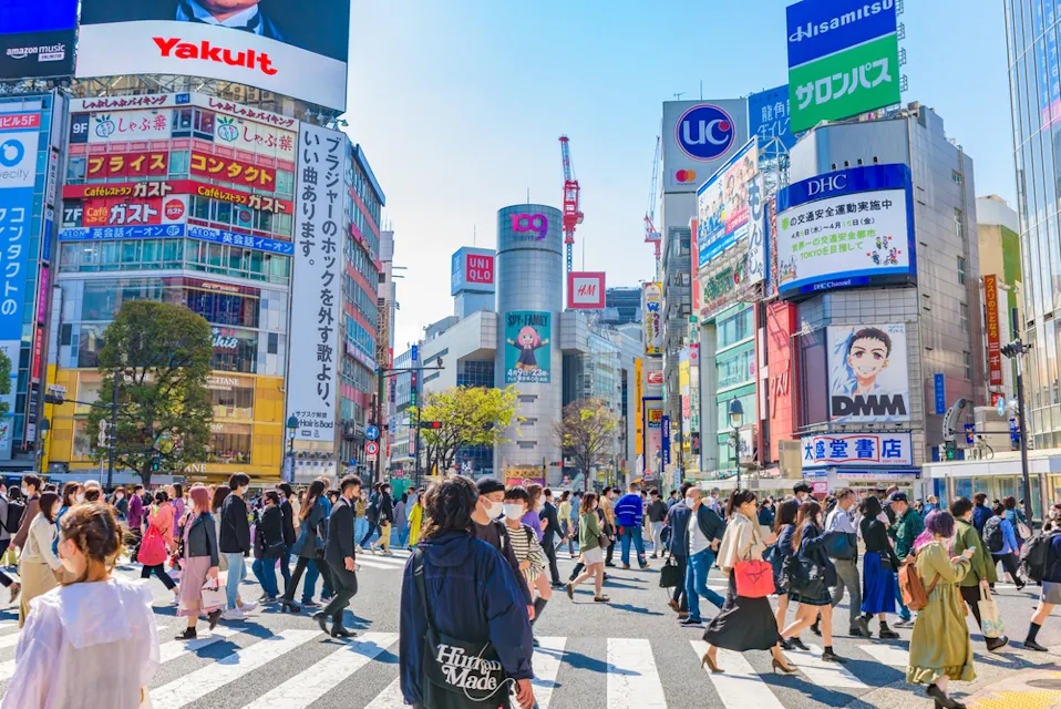 A bustling street scene in Shibuya, Tokyo, with crowds of people crossing at a busy intersection. Skyscrapers adorned with colorful advertisements and billboards surround the area under a clear blue sky.