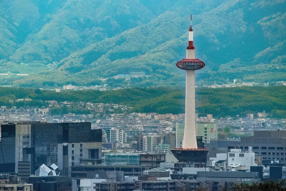 A modern cityscape featuring a tall tower with a red observation deck against a backdrop of lush green mountains under a clear blue sky. The city buildings are densely packed, hinting at a bustling urban environment.