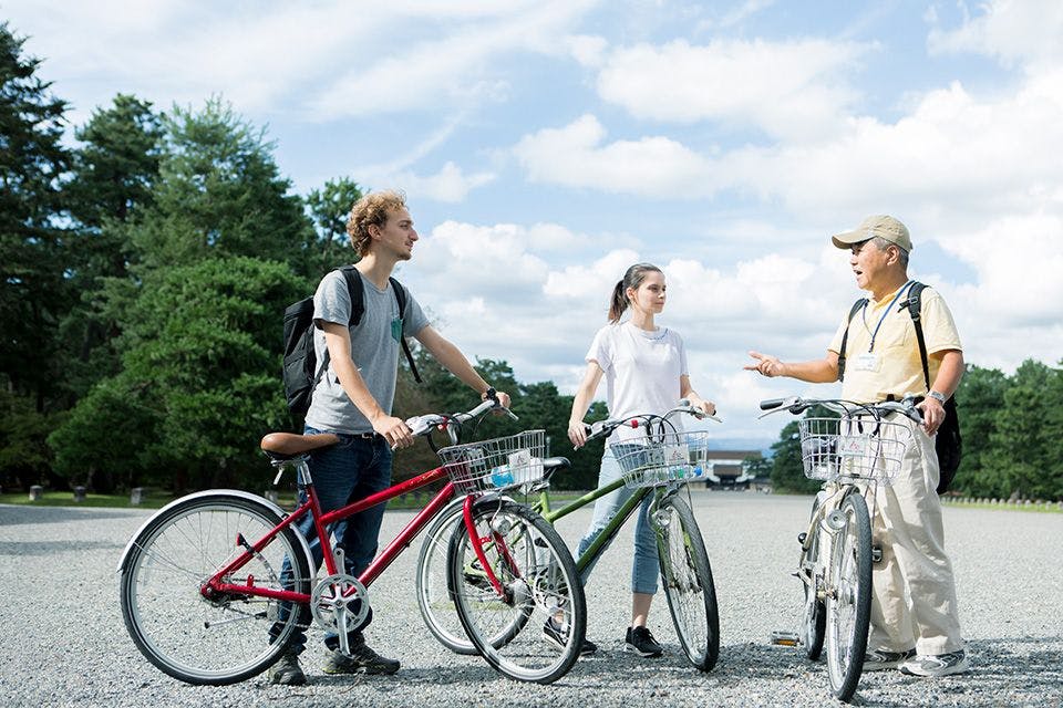 Three people with backpacks stand outdoors, each holding a bicycle and talking. They are on a gravel path with trees and a partly cloudy sky in the background. Two are young adults and one is older.