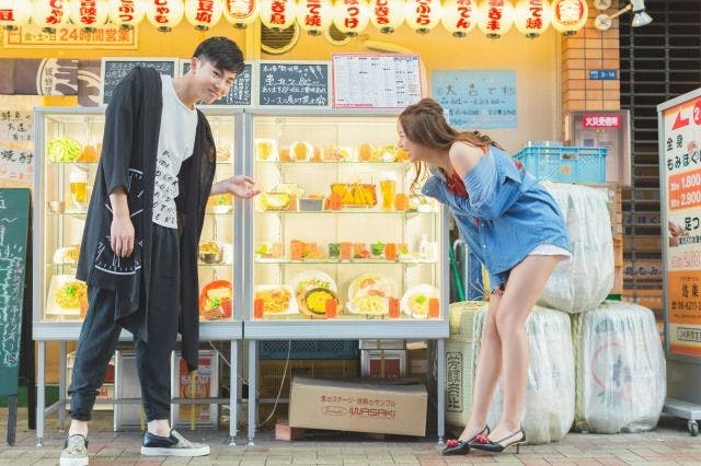 A man and woman stand outside a restaurant, looking and smiling at a display case of food models. Colorful lanterns and signs hang above, creating a lively street scene.