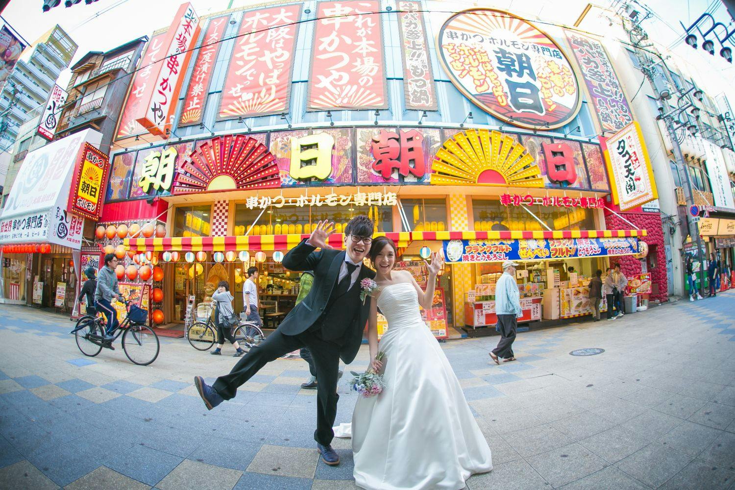 A joyful bride and groom pose playfully on a busy street in front of a colorful Japanese storefront with bright signs and lanterns, while pedestrians and cyclists pass by.