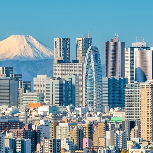 View of Mt. Fuji A city skyline with various tall buildings under a clear blue sky. A snow-capped mountain is prominently visible in the background, creating a striking contrast with the urban landscape.