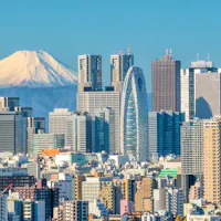 View of Mt. Fuji A city skyline with various tall buildings under a clear blue sky. A snow-capped mountain is prominently visible in the background, creating a striking contrast with the urban landscape.