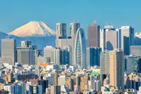 A city skyline with various tall buildings under a clear blue sky. A snow-capped mountain is prominently visible in the background, creating a striking contrast with the urban landscape.