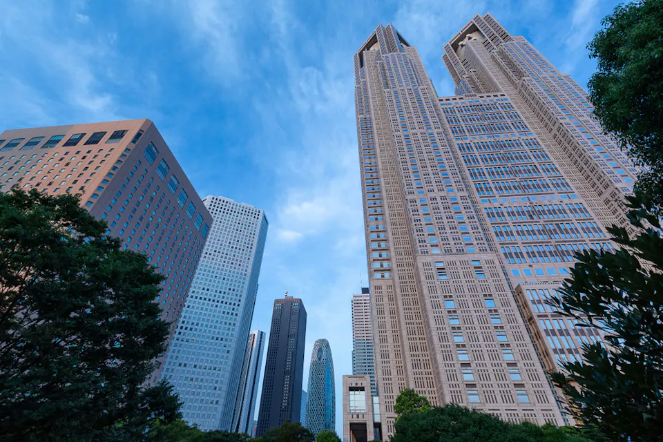A cityscape view featuring several modern skyscrapers, including a distinctive twin-tower building against a clear blue sky. Lush green trees are visible at the bottom, framing the urban skyline.