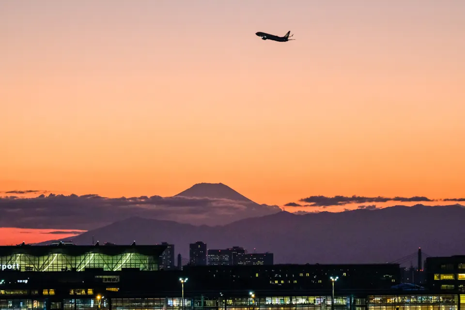 A silhouette of an airplane taking off against an orange and pink sunset sky, with Mount Fuji in the background. The foreground includes a building with bright lights, likely an airport terminal.