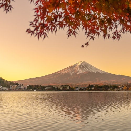 Lake Kawaguchiko A serene view of Mount Fuji with a snow-capped peak during sunset, framed by autumn leaves. The mountain reflects on a calm lake, with houses dotting the far shoreline under a warm, glowing sky.
