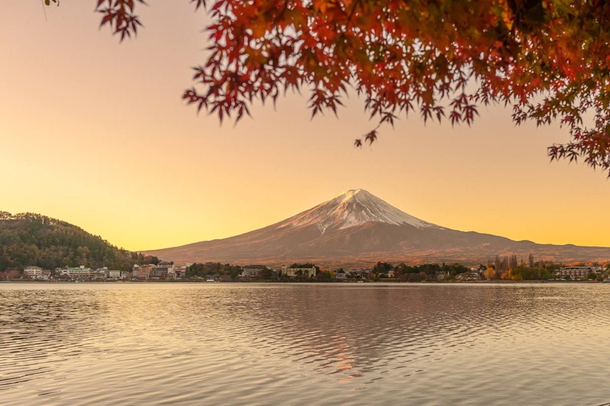 Lake Kawaguchiko A serene view of Mount Fuji with a snow-capped peak during sunset, framed by autumn leaves. The mountain reflects on a calm lake, with houses dotting the far shoreline under a warm, glowing sky.