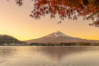 A serene view of Mount Fuji with a snow-capped peak during sunset, framed by autumn leaves. The mountain reflects on a calm lake, with houses dotting the far shoreline under a warm, glowing sky.