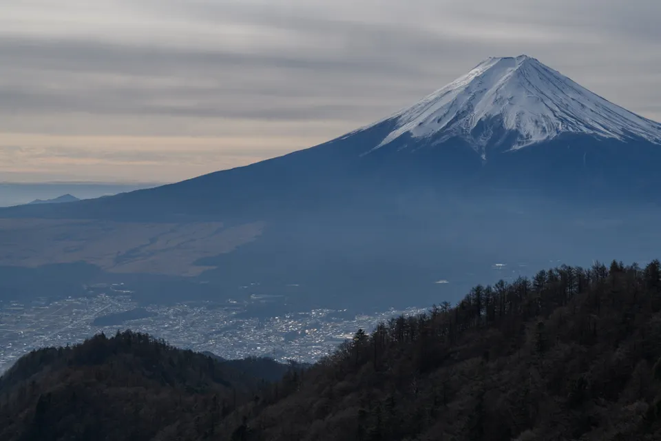 A majestic snow-capped mountain stands tall under a cloudy sky, overlooking a landscape of hills and a sprawling town nestled below. Dense forests cover the foreground mountains, adding depth to the scene.