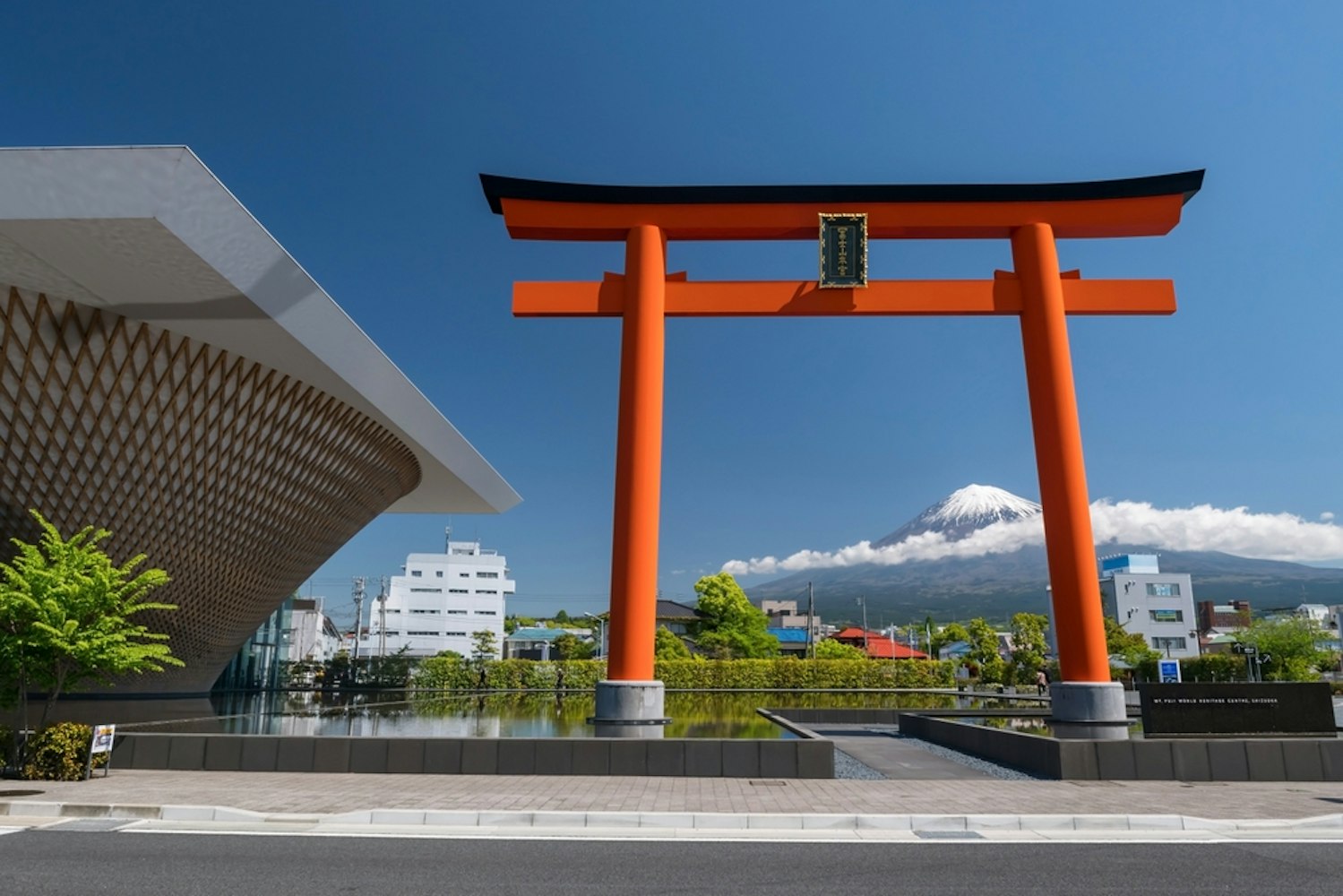 A large red torii gate stands prominently under a clear blue sky, with Mount Fuji visible in the background. Modern buildings and a uniquely designed structure are seen nearby, surrounded by greenery.