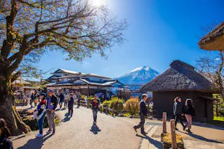 People stroll through a picturesque village with traditional Japanese architecture under a clear blue sky. Mount Fuji looms majestically in the background, framed by trees and vibrant sunlight.