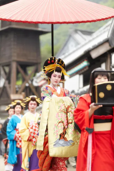 A woman in ornate traditional Japanese attire and elaborate hair ornaments walks under a large red parasol, leading a procession of similarly dressed women during a cultural parade. Traditional buildings are visible in the background.
